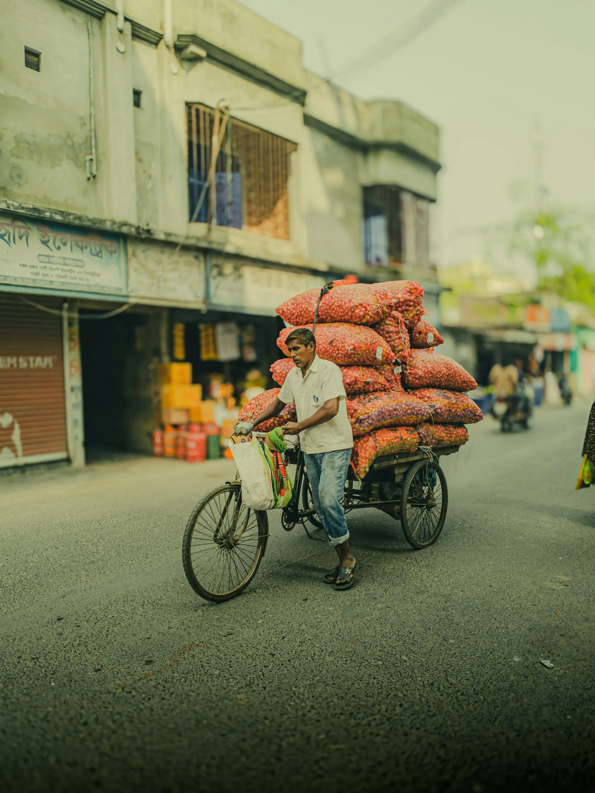 A man pedals a three-wheeled van loaded high with orange sacks down a quiet street lined with shopfronts.