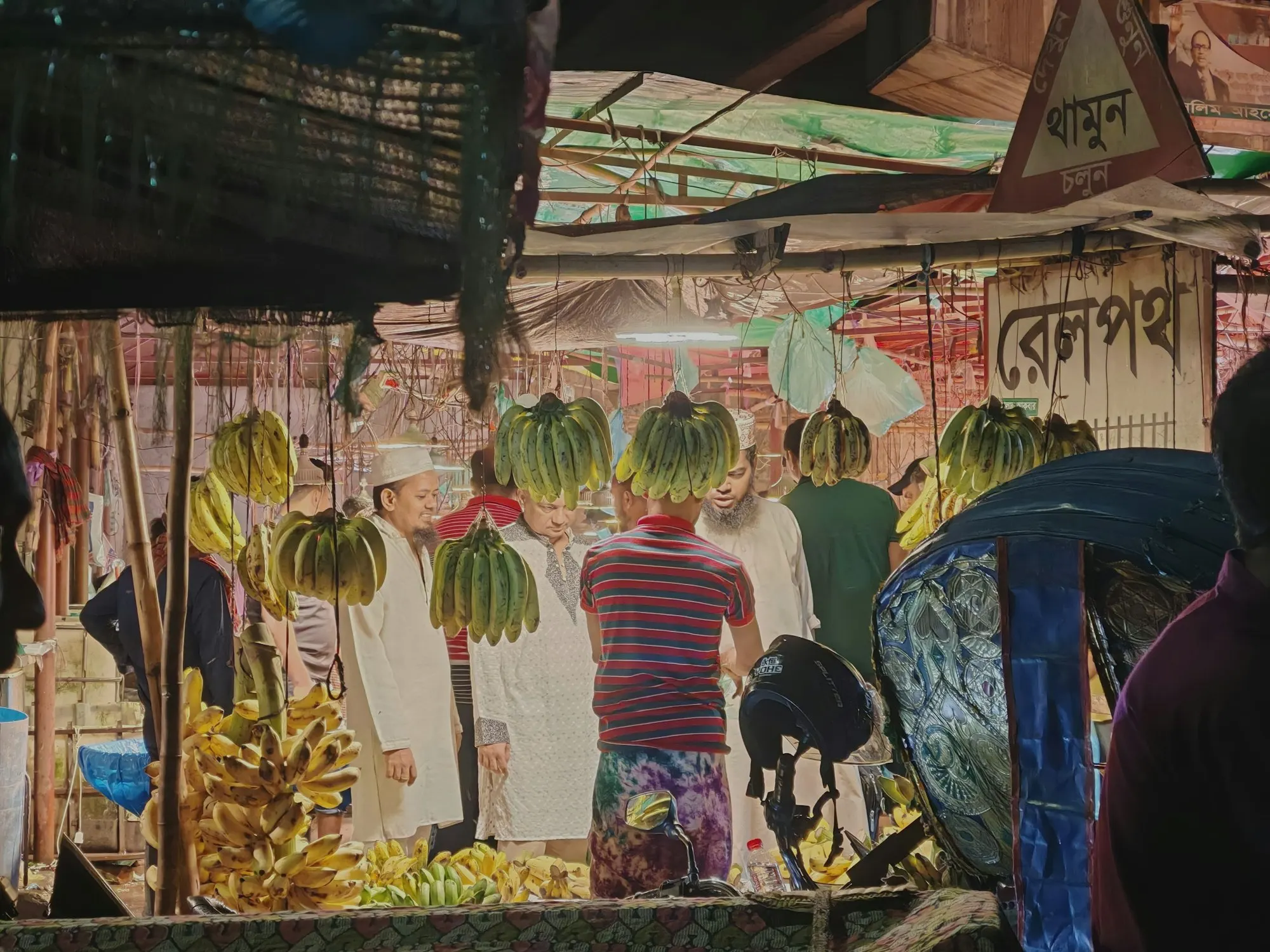 Bunches of green bananas hang over a market stall at night while shoppers browse beneath painted signs.