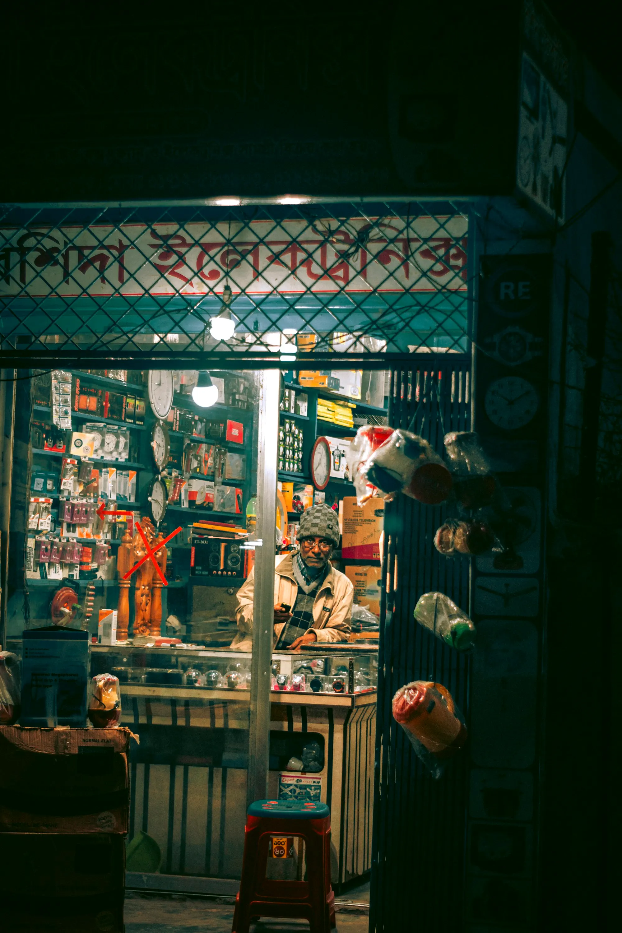 A small electronics shop at night, shelves packed with goods under a painted shop sign, the owner seated at the counter.